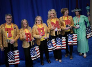 Apoiadores defendem Trump e guerra com Irã na CPAC – 26/03/2026 – Mundo Seis pessoas alinhadas em fundo azul, cinco vestem jaquetas douradas com letras vermelhas formando