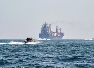 Irã força recuo de navios no estreito de Hormuz – 27/03/2026 – Mundo Navio de carga ao fundo no mar aberto com fumaça saindo da chaminé. Em primeiro plano, embarcação rápida com quatro pessoas navegando em direção ao navio.