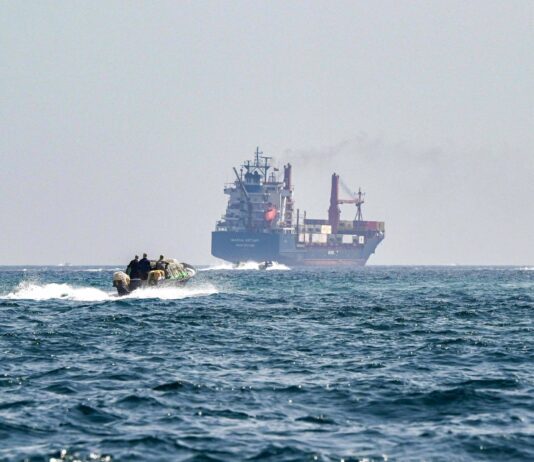 Irã força recuo de navios no estreito de Hormuz – 27/03/2026 – Mundo Navio de carga ao fundo no mar aberto com fumaça saindo da chaminé. Em primeiro plano, embarcação rápida com quatro pessoas navegando em direção ao navio.