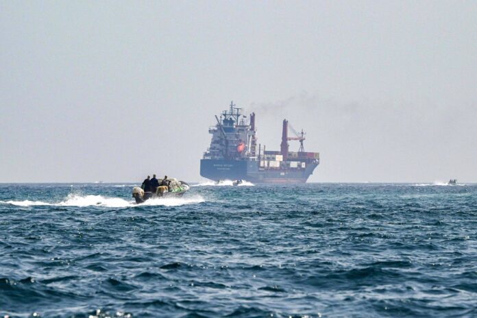 Navio de carga ao fundo no mar aberto com fumaça saindo da chaminé. Em primeiro plano, embarcação rápida com quatro pessoas navegando em direção ao navio.