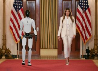 Melania Trump desfila com robô na Casa Branca – 26/03/2026 – Mundo Mulher vestindo terno branco caminha ao lado de robô humanoide branco e preto em salão com tapete vermelho. Duas bandeiras dos Estados Unidos estão posicionadas atrás deles, uma de cada lado, diante de portas de madeira.