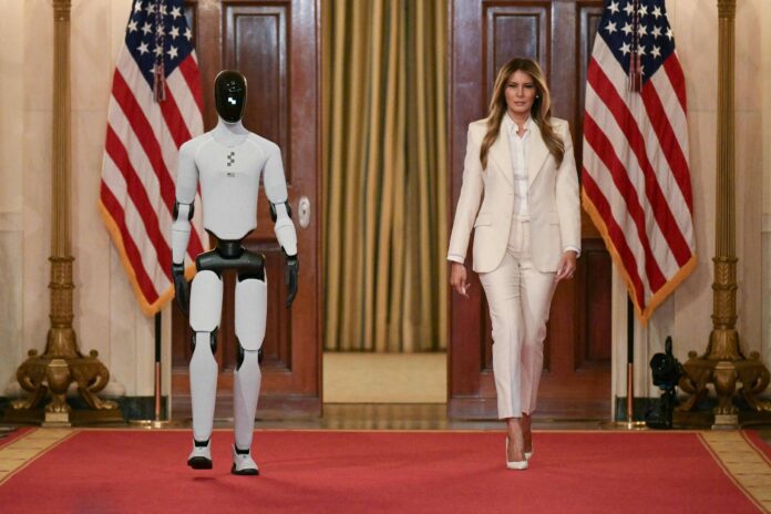 Mulher vestindo terno branco caminha ao lado de robô humanoide branco e preto em salão com tapete vermelho. Duas bandeiras dos Estados Unidos estão posicionadas atrás deles, uma de cada lado, diante de portas de madeira.
