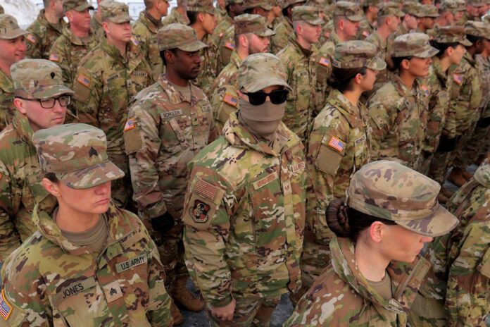 Grupo de soldados do Exército dos EUA em uniforme camuflado, alinhados em formação. Um soldado no centro usa óculos escuros e máscara facial, destacando-se entre os demais.