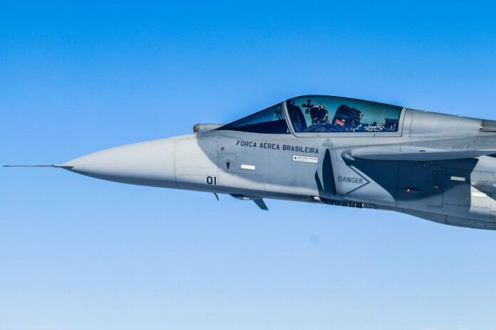 Aeronave de caça F-5 da Força Aérea Brasileira voa em céu claro. Piloto visível na cabine com capacete e equipamentos de voo.