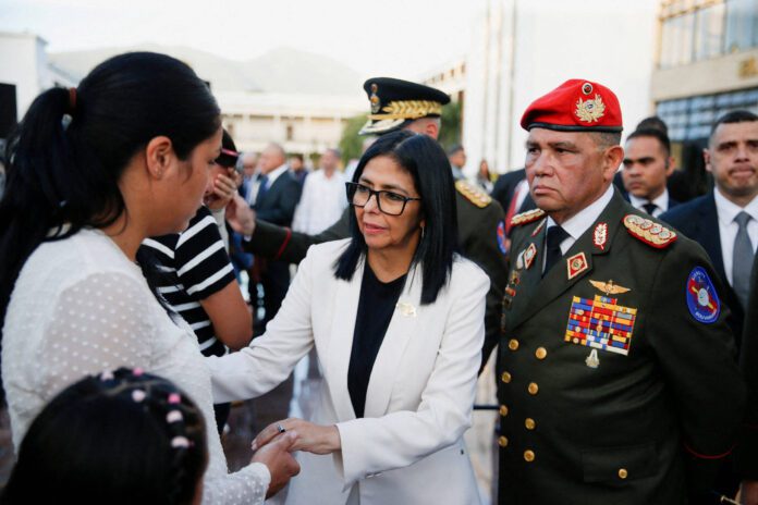 Mulher de terno branco e óculos cumprimenta outra mulher vestindo blusa branca com bolinhas, em ambiente externo com outras pessoas ao fundo. Homem em uniforme militar com boina vermelha observa ao lado.