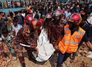 Vídeo: Ônibus cai em rio em Bangladesh e mata 24 pessoas – 26/03/2026 – Mundo Quatro socorristas com uniformes laranja e capacetes vermelhos carregam dois sacos brancos entre escombros. Multidão observa ao fundo próximo a corpo d