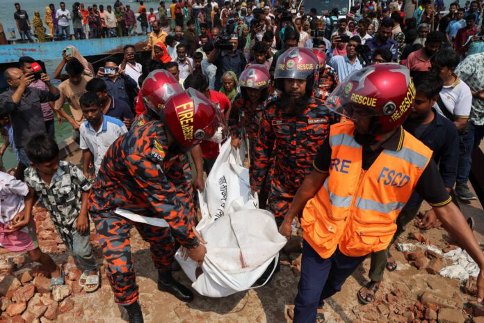 Vídeo: Ônibus cai em rio em Bangladesh e mata 24 Quatro socorristas com uniformes laranja e capacetes vermelhos carregam dois sacos brancos entre escombros. Multidão observa ao fundo próximo a corpo d